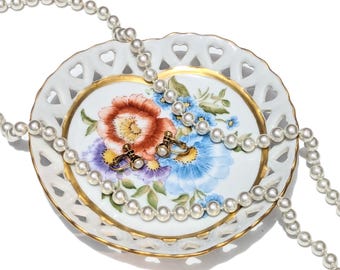 May include: A white pearl necklace and a pair of gold cufflinks with pearl accents rest on a decorative floral plate. The plate has a gold rim and heart-shaped cutouts. The plate's floral design features red, blue, and purple flowers.