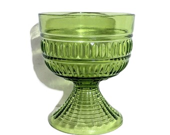 May include: A green glass goblet with a decorative design. The goblet has a wide bowl, a textured band around the middle, and a detailed stem. The glass has a vintage aesthetic and is set against a white background.