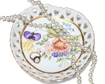 May include: A white, heart-shaped, decorative dish with a gold rim, featuring a floral design and a gold ring with a pearl. A pearl necklace is draped across the dish.