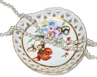 May include: A white, heart-shaped porcelain trinket dish with a gold rim, floral design, and openwork detailing. Adorned with a pearl necklace, earrings, and a ring. A classic jewelry organizer.