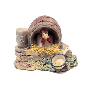 May include: A detailed figurine of a hen inside a barrel-shaped coop. The hen has a white body, red comb, and yellow beak. A small pan with yellow food and a pillar with the letter "B" are also present. The figurine is on a base of straw and wood.