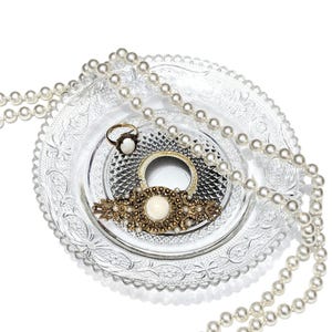 May include: An arrangement of vintage jewelry on a clear glass plate. The jewelry includes a pearl necklace, a gold-toned ring, a gold-toned bracelet, and a decorative brooch with a large white stone. The plate has an ornate, embossed design.