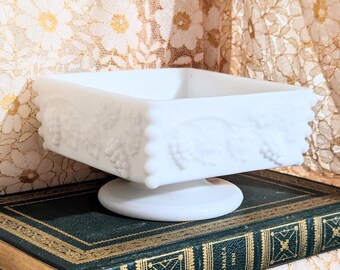 May include: A white, square, pedestal dish with a grape vine design. The dish is made of opaque glass and sits on a round base. The dish is resting on a vintage book with gold lettering.