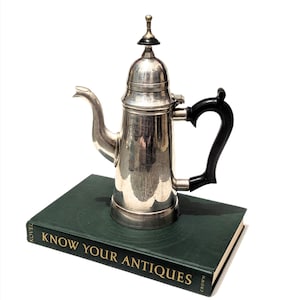 May include: A silver-colored antique coffee pot with a black handle and a decorative top, resting on a green book. The book's title, "KNOW YOUR ANTIQUES," is visible in gold lettering. The pot has a spout and a lid.