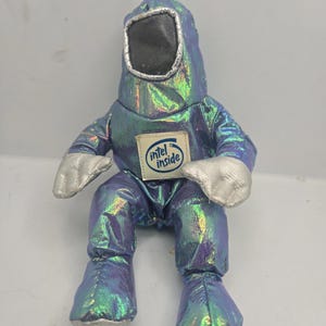 Puede incluir: Un peluche con forma de traje de protección, con una tela brillante e iridiscente azul-verdosa. El juguete tiene guantes plateados y una visera con borde plateado. Un parche de "Intel Inside" está cosido en el pecho.