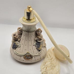 May include: A vintage perfume bottle with a cream-colored base and gold accents. Two cherubic figures flank the bottle, holding grapes. The bottle has a cream-colored bulb atomizer with a tassel.