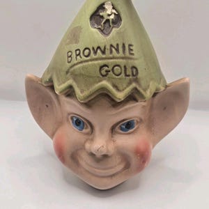 Könnte beinhalten: Ein Keramikgefäß in Form eines Elfenkopfes mit einer spitzen grünen Mütze, die den Schriftzug BROWNIE GOLD trägt. Der Elf hat große Ohren, blaue Augen und rosa Wangen. Die Mütze hat ein kleines dunkles Emblem mit einer hellen Figur darauf.