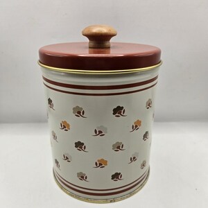 May include: A cylindrical metal canister with a cream-colored body featuring a repeating pattern of small brown, orange, and gray flowers. The lid is dark red with a gold rim and a central wooden knob handle. Two dark red horizontal stripes.