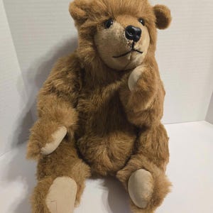 May include: A brown teddy bear with a soft, plush fur texture. The bear has a black nose, small black eyes, and tan-colored paws. It is sitting upright with one paw raised, giving it a thoughtful appearance. A classic children's toy.
