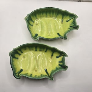 Peut inclure: Deux plats en céramique en forme de cochon. Les plats sont de couleur jaune clair avec une bordure vert foncé. Les plats ont une finition brillante et sont probablement faits à la main. Les plats sont un article unique et décoratif.
