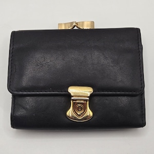 May include: A black leather wallet featuring a gold-toned metal clasp closure on the front flap and a gold-toned kiss-lock frame closure at the top. The wallet has a smooth, matte finish and a rectangular shape with rounded corners.