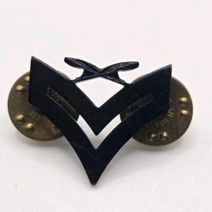 May include: Black metal military insignia with two chevrons and a crossed design on top. The insignia is attached to two gold-colored pin backs. The pin is likely a rank insignia.