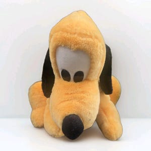 Pode incluir: Um brinquedo de pelúcia de um cão amarelo com orelhas pretas compridas, um nariz preto e olhos grandes e ovais. O personagem tem uma textura macia e felpuda e é retratado em uma posição sentada com as patas dianteiras voltadas para a frente.