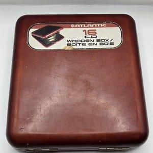 May include: A dark brown wooden CD storage case with rounded corners and metal hinges. A rectangular label on the top surface features the Atlantic logo, the number 16, and the text "16 CD WOODEN BOX/BOÎTE EN BOIS" with a small image of the open case.