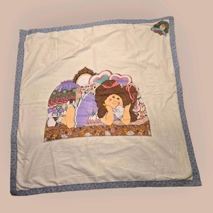 Pode incluir: Um edredom creme com uma borda floral azul. O centro apresenta um gráfico colorido de uma boneca com cabelo castanho, um vestido roxo e um bicho de pelúcia branco. A boneca está deitada. O edredom é quadrado.