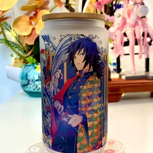Op de afbeelding: Een witte keramische beker met een bamboe deksel en een rietje. De beker heeft een kleurrijke illustratie van een personage uit de anime Demon Slayer. Het personage draagt een blauw-groene gepatroneerde kimono en houdt een zwaard vast. De achtergrond van de illustratie is een blauw-wit golfpatroon.