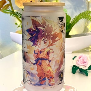 Op de afbeelding: Een frosted glazen beker met een bamboe deksel en een rietje. De beker heeft een kleurrijke illustratie van een cartoonfiguur met stekelig oranje haar, die een oranje jumpsuit en een blauwe riem draagt. Het figuur staat voor een gele en oranje achtergrond met zwart-wit manga-stijl illustraties.