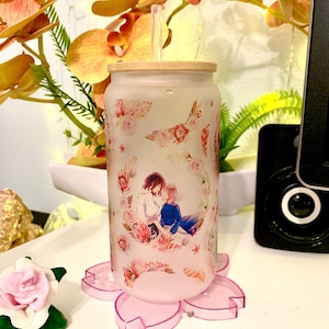 Op de afbeelding: Een frosted glazen beker met een bamboe deksel en een rietje. De beker heeft een bloemenpatroon met een stel in het midden. De beker staat op een roze bloemachtige onderzetter.
