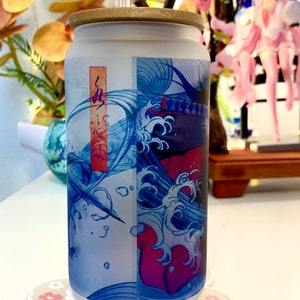 Op de afbeelding: Een matglazen beker met een bamboe deksel en een rietje. De beker heeft een Japans ge&iuml;nspireerd ontwerp in blauw en rood met golven en een figuur in het midden. De beker staat op een witte onderzetter met roze bloemen.