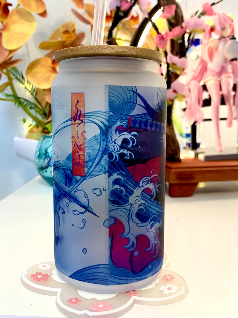 Op de afbeelding: Een matglazen beker met een bamboe deksel en een rietje. De beker heeft een Japans ge&iuml;nspireerd ontwerp in blauw en rood met golven en een figuur in het midden. De beker staat op een witte onderzetter met roze bloemen.