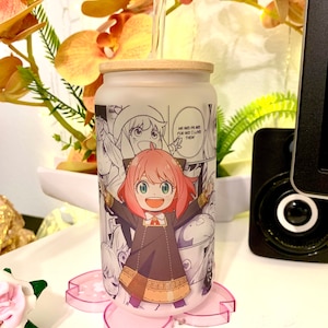 Op de afbeelding: Een witte herbruikbare glazen beker met een roze en wit anime-personageontwerp. De beker heeft een bamboe deksel en een doorzichtige plastic rietje. De beker staat op een roze bloemachtige onderzetter.