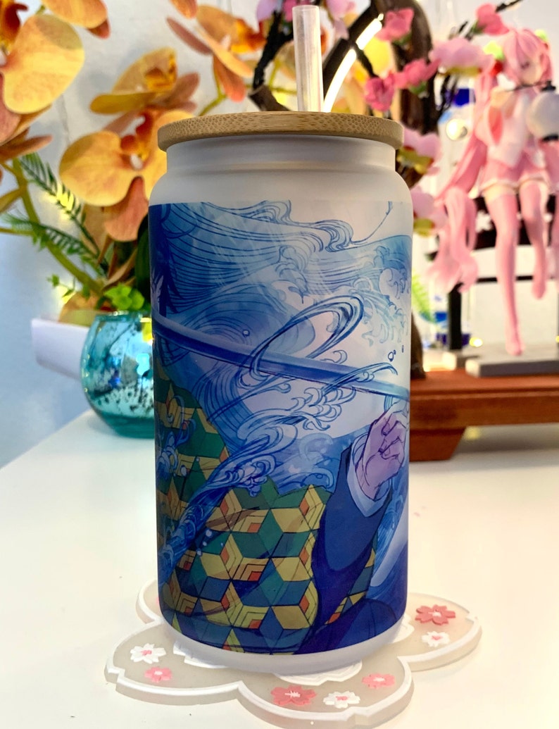 Op de afbeelding: Een witte tumbler met een blauw en groen ontwerp met een personage uit de anime Demon Slayer. De tumbler heeft een bamboe deksel en een rietje.