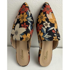 May include: A pair of floral print slip-on shoes. The shoes feature a black background with a pattern of orange, yellow, and white flowers. The brand name "Tilocow" is printed in gold on the insole. The shoes have pointed toes.
