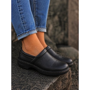 May include: A pair of black leather slip-on clogs with a textured grain finish, a rounded toe, and a low heel. The shoes feature visible stitching along the sole and a small embossed logo on the side, paired with blue denim jeans.