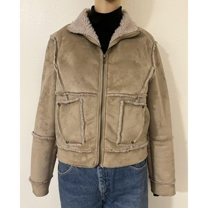 May include: A tan suede jacket with a faux fur collar and trim. The jacket has a zip-up front, two front pockets, and long sleeves. The model is wearing a black turtleneck and blue jeans.