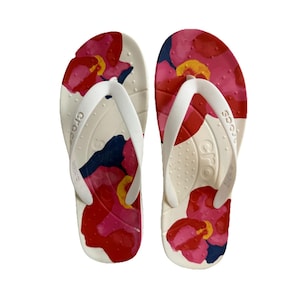 May include: A pair of white Crocs flip-flops with a floral pattern. The flip-flops feature a cream-colored base with a vibrant design of red, pink, yellow, and navy blue flowers. The straps are white.