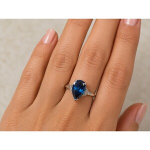 May include: A silver-toned ring featuring a large, pear-shaped, deep blue gemstone as the centerpiece. The band is accented with small, clear, sparkling stones on either side of the central setting, creating a refined and polished appearance.