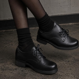 May include: A pair of black leather oxford shoes with matching black laces, a rounded toe, and a low block heel. The shoes are worn with black ribbed crew socks and sheer black tights, showcasing a classic lace-up design and smooth finish.