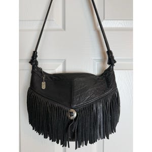 May include: A black leather shoulder bag featuring a long fringe trim along the bottom edge, a single thin shoulder strap with knotted detailing at the sides, and a circular silver-toned metal concho ornament centered on the front panel.