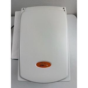 May include: A light grey flatbed scanner with a rectangular body and rounded corners. An orange oval emblem is positioned near the bottom centre, featuring the text "Mustek" and "1200 UB" printed in white lettering on the surface of the device.