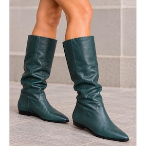 Könnte beinhalten: Ein Paar kniehohe Lederstiefel in Dunkel-Türkis mit lockerem Schaft, spitzer Zehenpartie und flacher schwarzer Sohle. Die Stiefel haben eine glatte Oberfläche, reichen bis knapp unter das Knie und zeigen eine strukturierte, aber lockere Form.