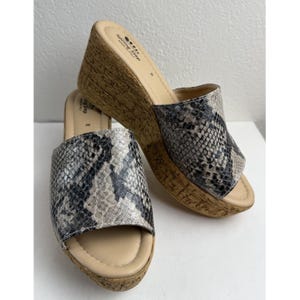Op de afbeelding: Een paar Spring Step sleehak sandalen. De sandalen hebben een slangenprint in grijstinten, zwart en beige. De sleehakken hebben een kurkachtige textuur en de voetbedden zijn lichtbeige. De sandalen zijn ontworpen voor comfort en stijl.