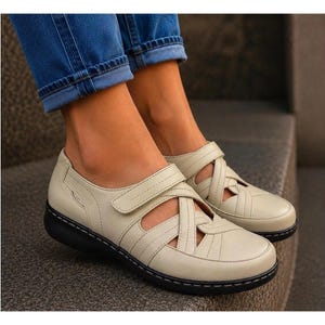 May include: Off-white leather shoes featuring a crisscross strap design across the top, a hook-and-loop closure, a rounded toe, and a thick black sole with visible stitching along the edge, paired with blue denim jeans.