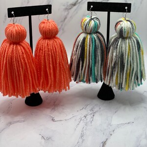 May include: Two pairs of colorful yarn tassel earrings. One pair is a bright orange color and the other pair is a multi-colored striped pattern.
