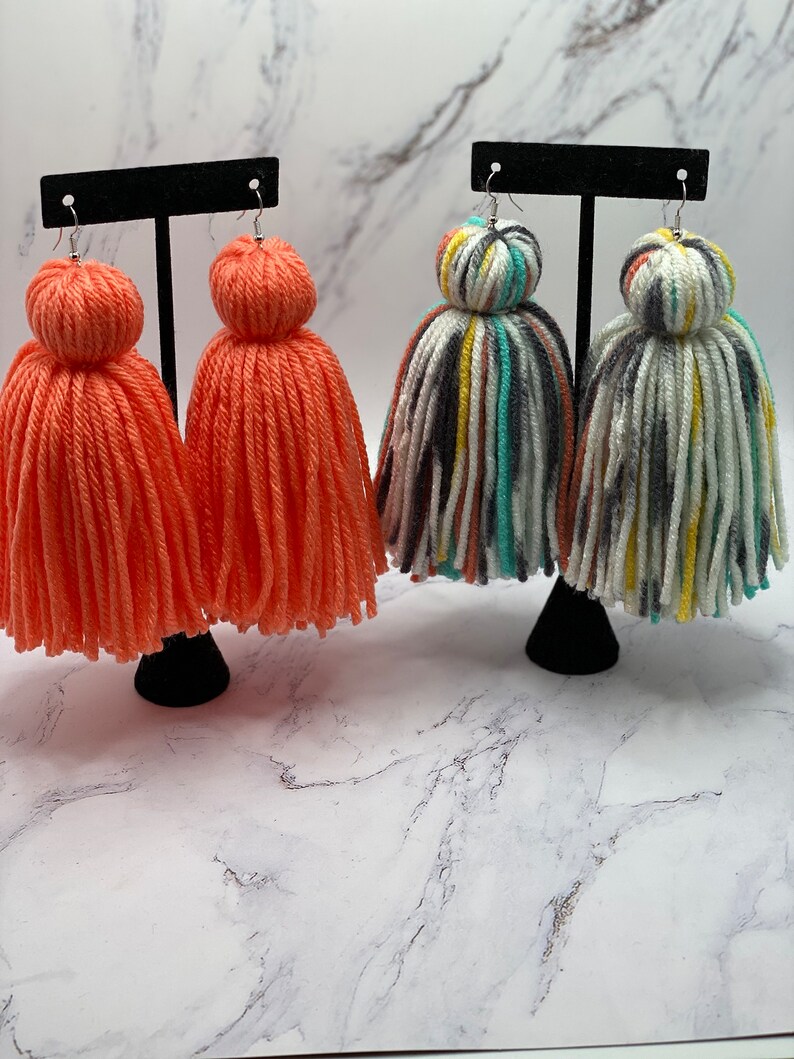 May include: Two pairs of colorful yarn tassel earrings. One pair is a bright orange color and the other pair is a multi-colored striped pattern.