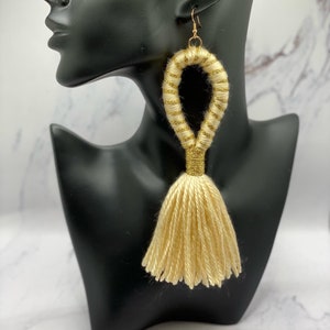 Op de afbeelding: Gouden oorhangers met kwastjes. De oorbellen hebben een lusontwerp met gouden glitteraccenten en een lange kwast. De oorbellen worden getoond op een zwarte mannequinhoofd tegen een marmeren achtergrond.