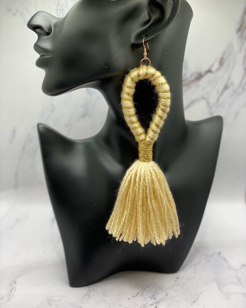 Op de afbeelding: Gouden oorhangers met kwastjes. De oorbellen hebben een lusontwerp met gouden glitteraccenten en een lange kwast. De oorbellen worden getoond op een zwarte mannequinhoofd tegen een marmeren achtergrond.