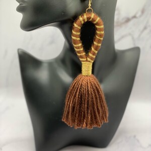 Op de afbeelding: Bruine oorbel met kwastjes en gouden accenten. De oorbellen hebben een groot, lusvormig ontwerp dat is gewikkeld in bruin garen met gouden strepen. De onderkant van de oorbellen heeft een bruine kwast. De oorbellen worden getoond op een zwarte mannequin hoofd.