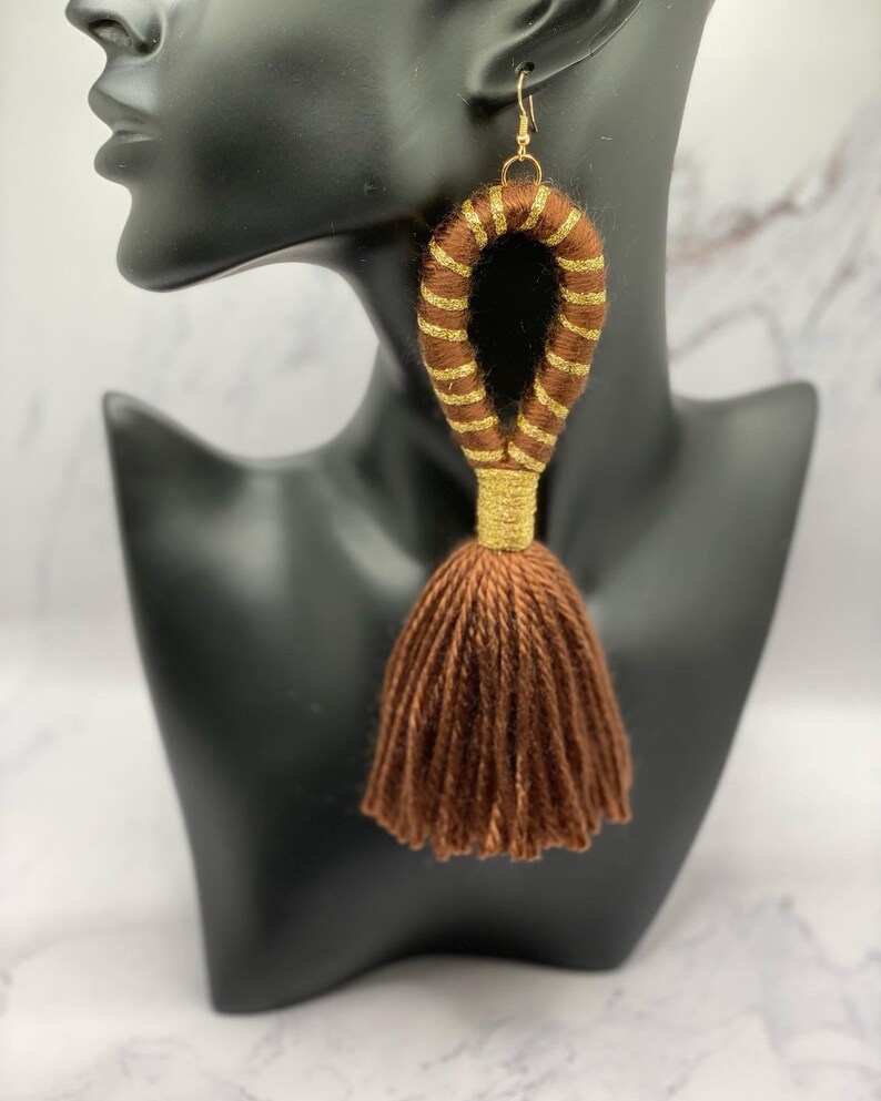 Op de afbeelding: Bruine oorbel met kwastjes en gouden accenten. De oorbellen hebben een groot, lusvormig ontwerp dat is gewikkeld in bruin garen met gouden strepen. De onderkant van de oorbellen heeft een bruine kwast. De oorbellen worden getoond op een zwarte mannequin hoofd.