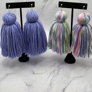 May include: Two pairs of yarn tassel earrings. One pair is a solid lavender color and the other pair is a multi-colored yarn with pink, green, white, and blue stripes.