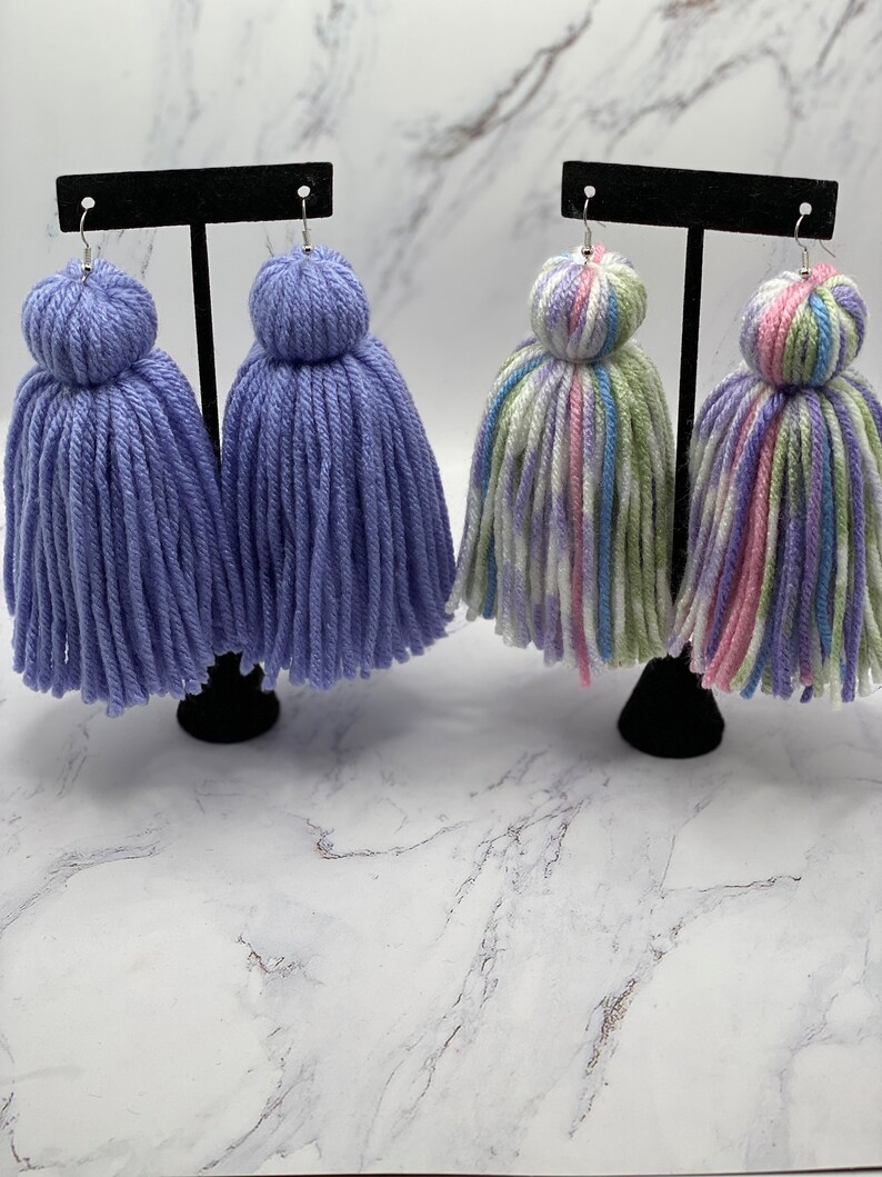 May include: Two pairs of yarn tassel earrings. One pair is a solid lavender color and the other pair is a multi-colored yarn with pink, green, white, and blue stripes.