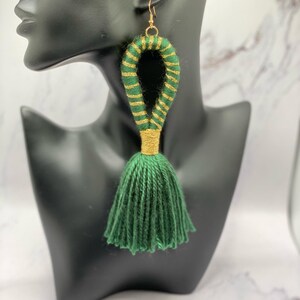 Op de afbeelding: Groene kwast oorbellen met goudkleurige accenten. De oorbellen hebben een lusontwerp met groen en goudkleurig garen en een groene kwast. De oorbellen worden gepresenteerd op een zwarte mannequinhoofd.