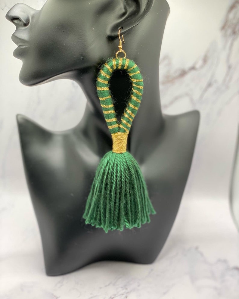 Op de afbeelding: Groene kwast oorbellen met goudkleurige accenten. De oorbellen hebben een lusontwerp met groen en goudkleurig garen en een groene kwast. De oorbellen worden gepresenteerd op een zwarte mannequinhoofd.