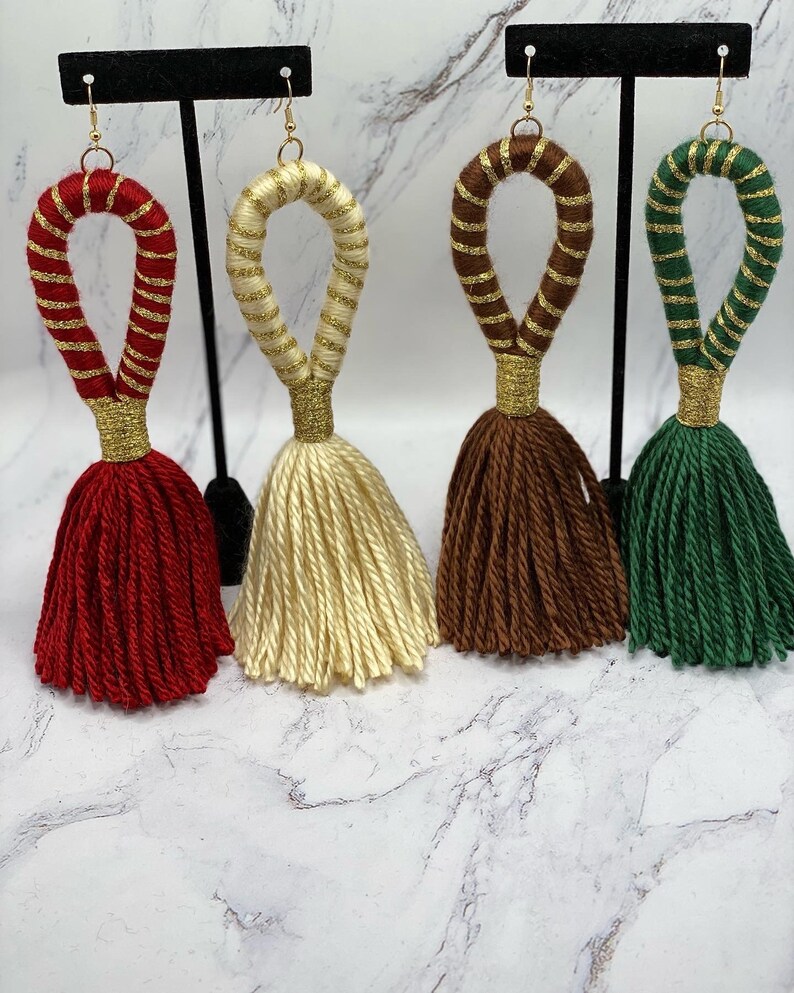 Op de afbeelding: Vier paar kwast oorbellen in rood, cr&egrave;me, bruin en groen. Elke oorbel heeft een lus gewikkeld met bijpassend garen en gouddraad, verbonden met een kwast. De oorbellen hangen aan goudkleurige haken.