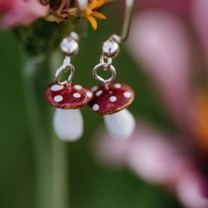 May include: A pair of silver mushroom earrings with red and white accents. The earrings are hanging from a plant.