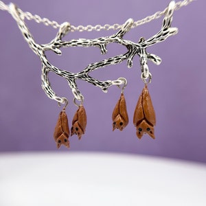 Op de afbeelding: Een zilveren takketingsketting met vier bruine vleermuisbedels die eraan hangen. De vleermuizen hangen naar beneden en hebben zwarte ogen.